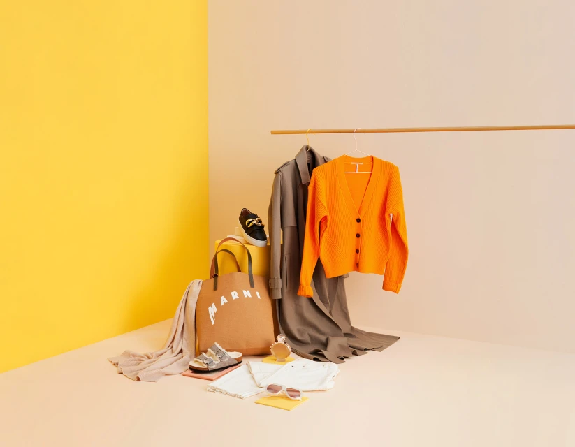 Fashion flat lay with an orange cardigan, tote bag, shoes, sunglasses, and other accessories against a yellow and beige studio backdrop.