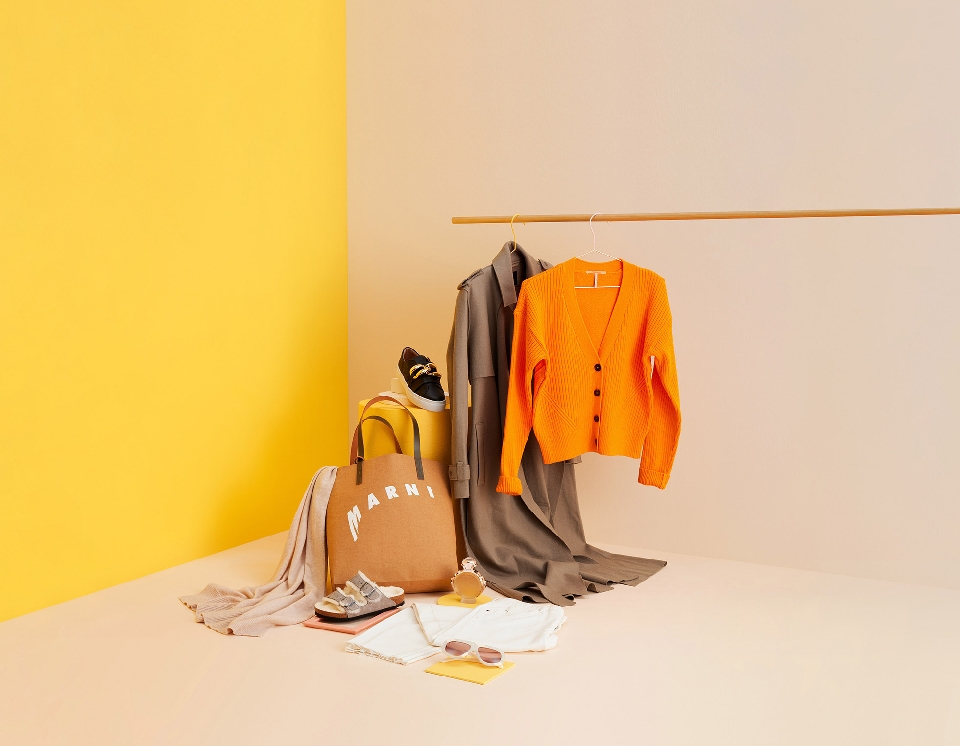 Fashion still life on peach and yellow background with a trench coat, orange cardigan, handbag, sandals, sneakers, and sunglasses.