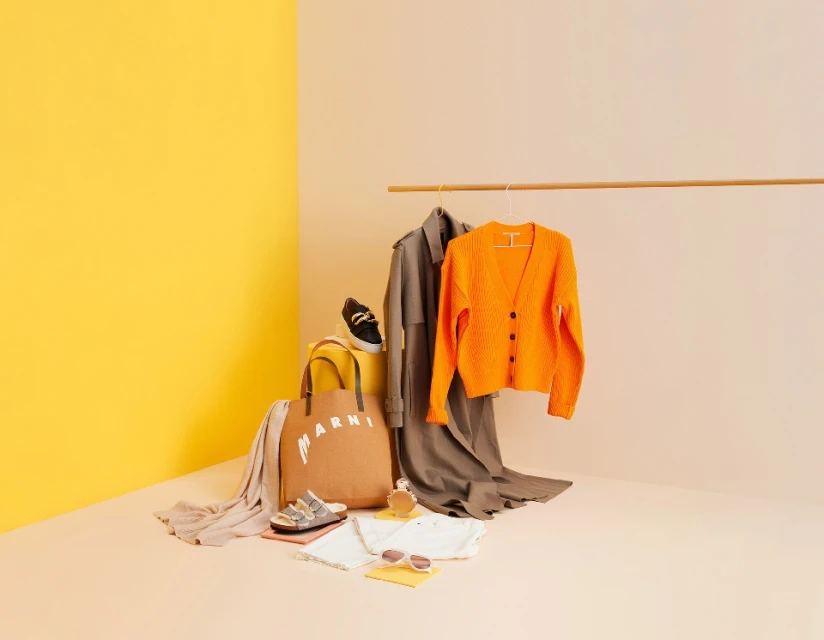 Fashion still life on peach and yellow background with a trench coat, orange cardigan, handbag, sandals, sneakers, and sunglasses.