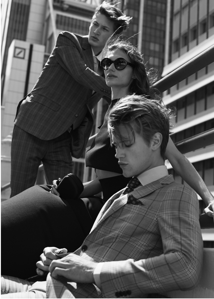 Monochrome fashion shot of three young adults outdoors with modern architecture. Two men in suits, one woman in sunglasses.