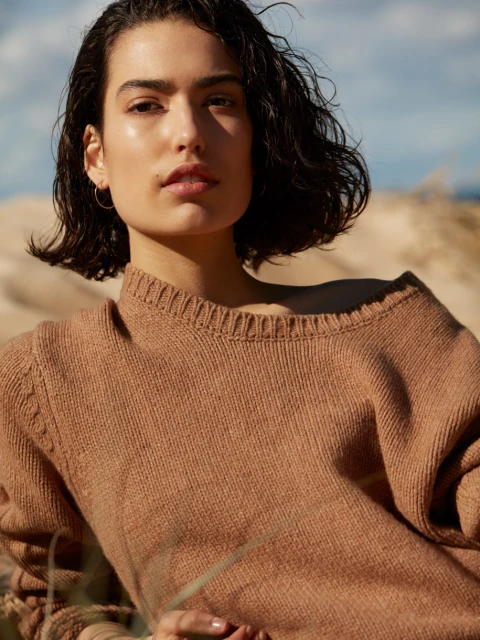 Portrait of an adult female model wearing a tan sweater, with short dark hair and minimalist jewelry, set against a sandy background.