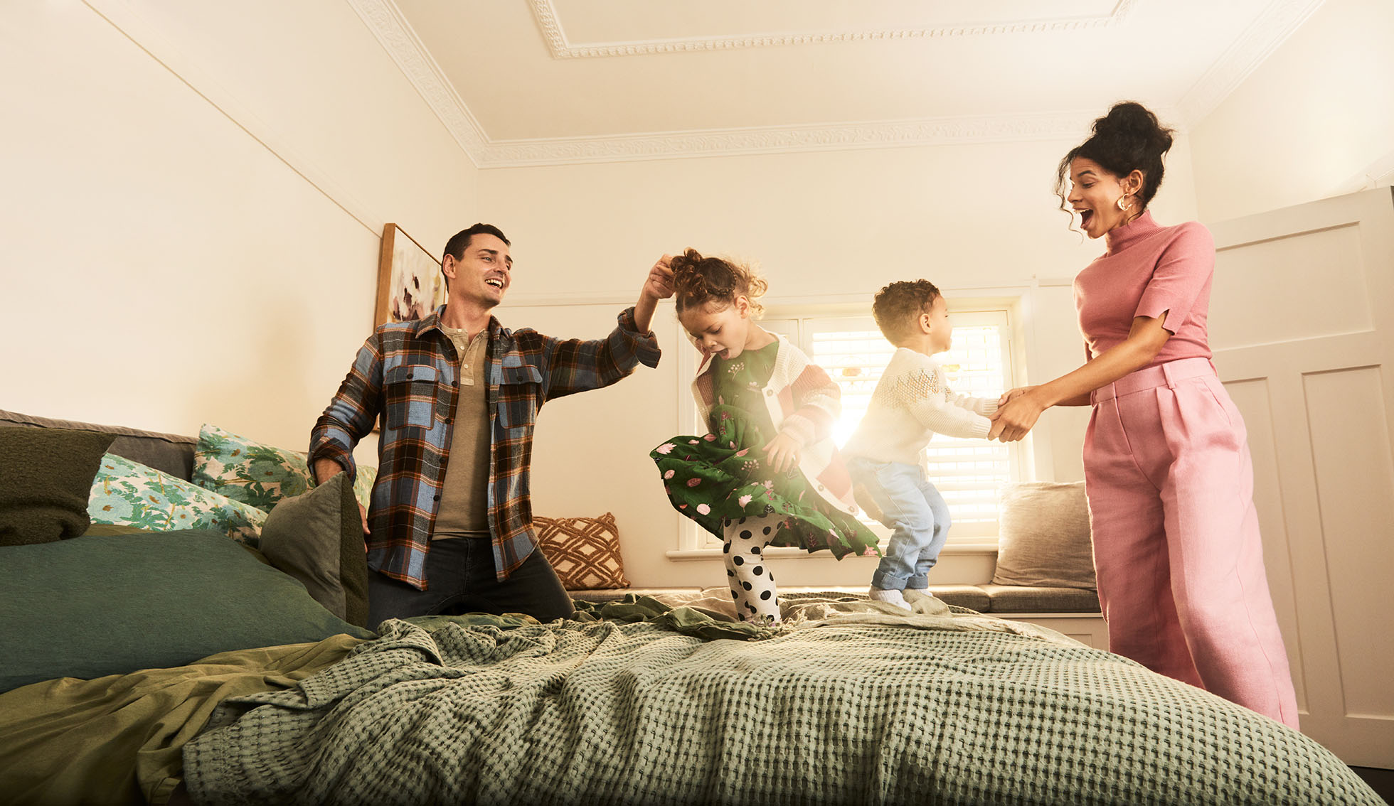 Family of four: father, mother, daughter, and son playing together on a bed in a bright, airy indoor setting with a window.