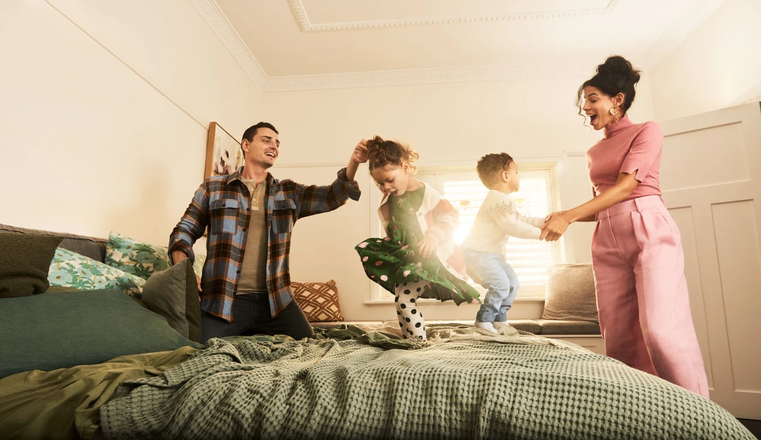 Family of four: father, mother, daughter, and son playing together on a bed in a bright, airy indoor setting with a window.