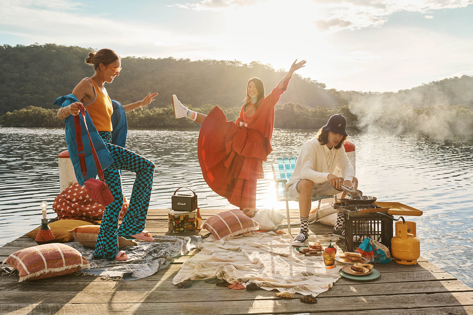 Three women and a man enjoying an outdoor picnic on a wooden deck by a lake with hills and trees in the background on a sunny day.