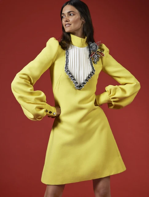 An adult female fashion model poses in a yellow minidress with a unique neck embellishment against a red backdrop, studio lit.