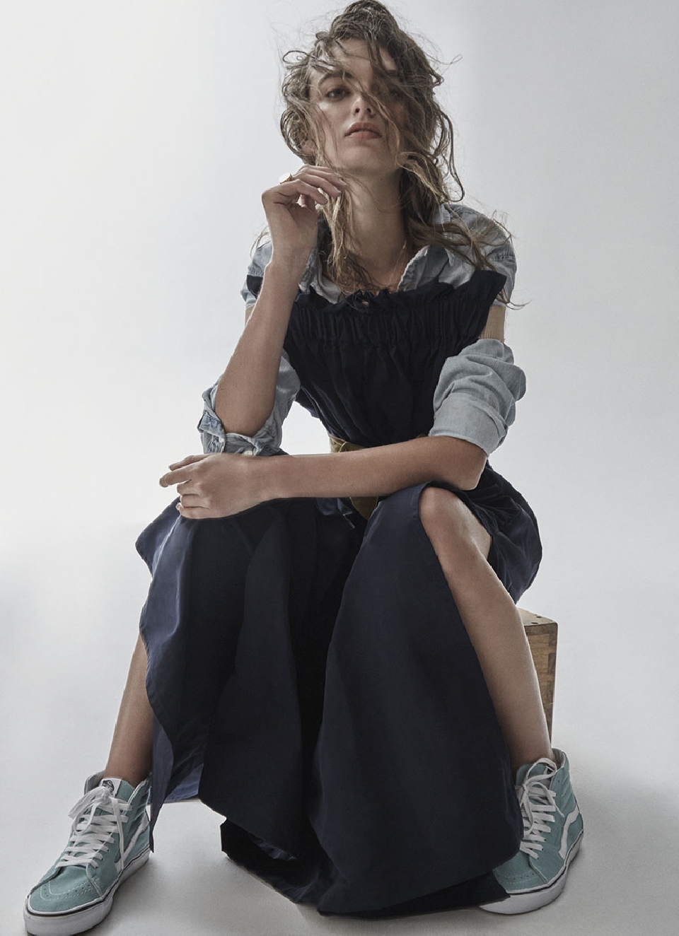 A young woman with brown hair sits on a wood block wearing a blue dress and sneakers, with a grey background, captured in a studio shot.