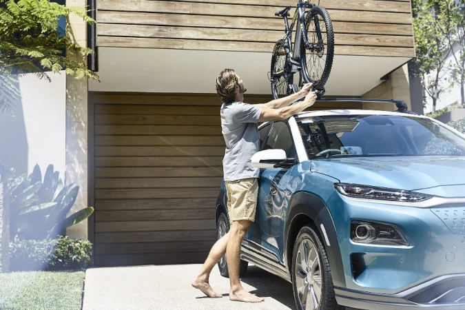 A fair-skinned adult male is placing a mountain bike onto a car's roof rack outside of a modern residence, showcasing an outdoor lifestyle scene.