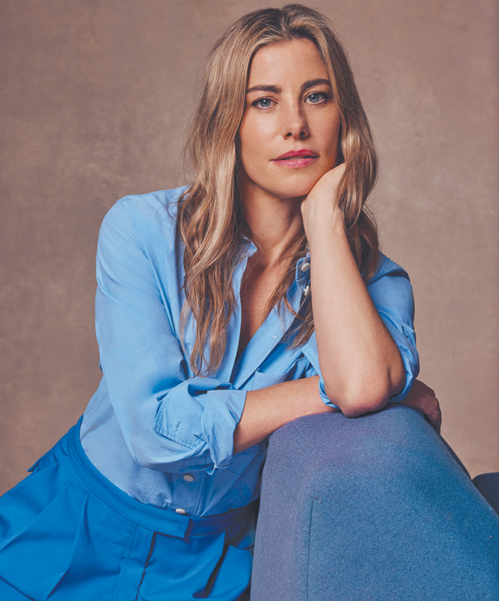 Portrait of a blonde woman in blue shirt and pants, leaning on a blue upholstered surface against a neutral backdrop in a photo studio.