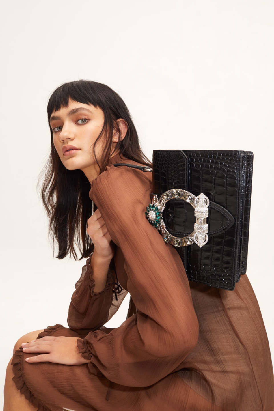 A stylish woman with dark hair poses indoors with a black crocodile-patterned clutch featuring a jeweled buckle detail, against a plain background.