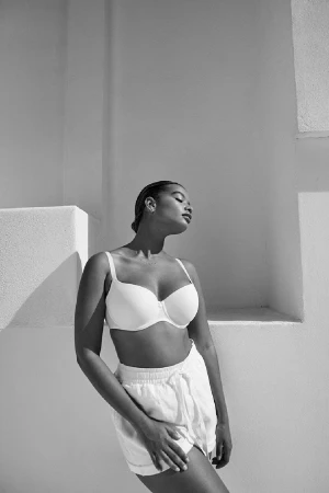 Black and white portrait of a woman wearing a white bra and shorts posing against a light architectural background.