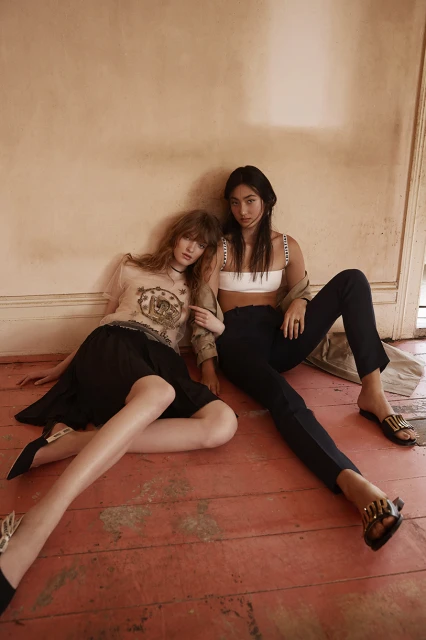 Two women sit indoors on a red-painted wooden floor against a peach wall. One woman wears a white crop top and trousers, the other a patterned top and skirt.