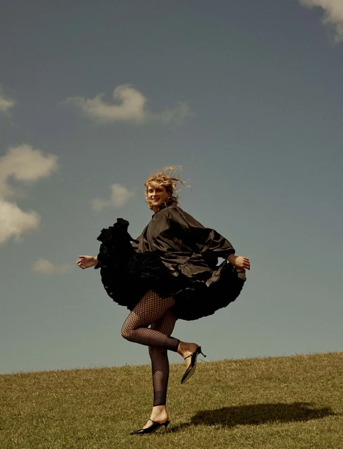 Woman in black dress and fishnet tights poses on grassy hill under blue sky with clouds in outdoor fashion shot.