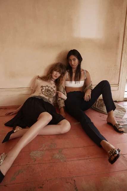 Two fashion models, one blonde and one with black hair, pose on a distressed wooden floor in black outfits with a neutral background.