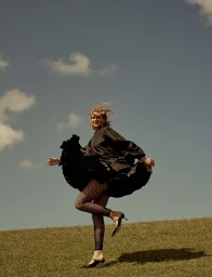 Smiling woman in black dress and fishnet tights jumping on a grassy hill under a blue sky, styled by Jackie Shaw.