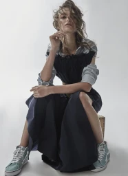 Model seated on a wooden crate wearing a dark dress over a striped shirt and light blue high top sneakers against a seamless white backdrop.