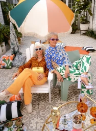 Two older women in patterned shirts and sunglasses sitting outdoors under a large colorful fringed umbrella styled by Jackie Shaw