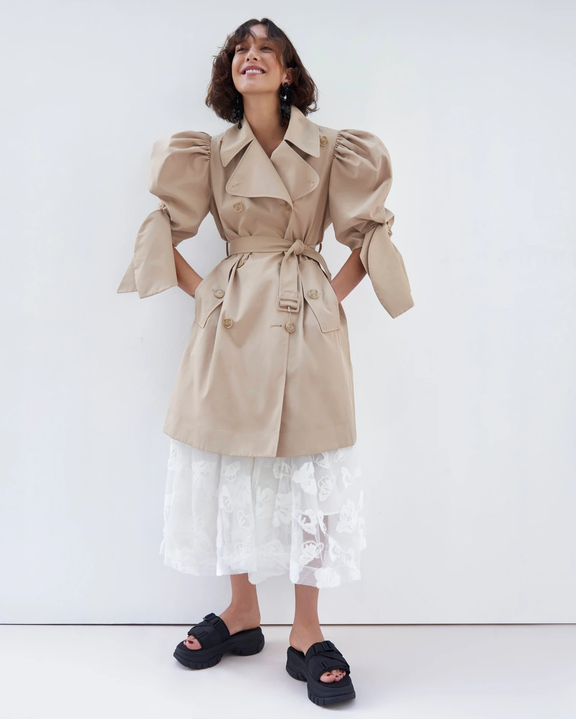 Model in beige belted trench coat with puff sleeves and white skirt standing against white backdrop