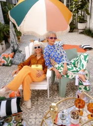 Two older women in patterned shirts and sunglasses relaxing outdoors under a colourful parasol styled by Jackie Shaw.