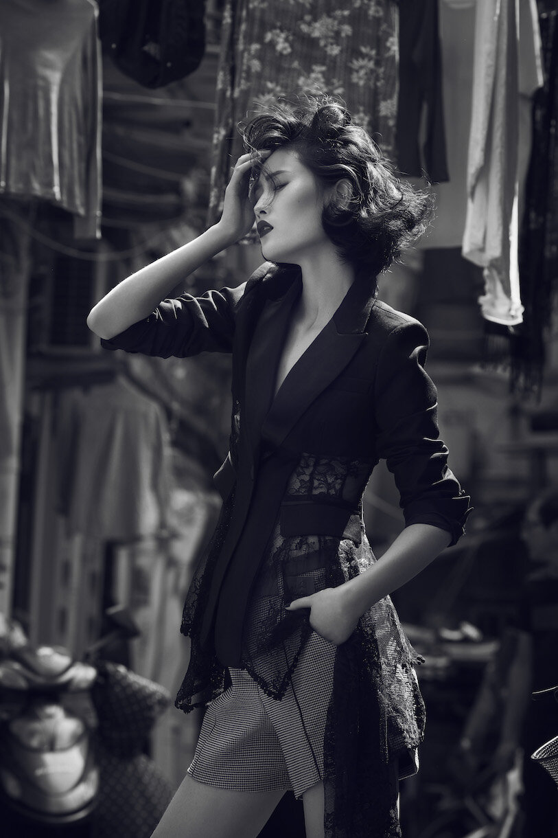 Monochrome fashion shot of a woman with short hair in a jacket and shorts posing in front of hanging clothes, dramatic style.