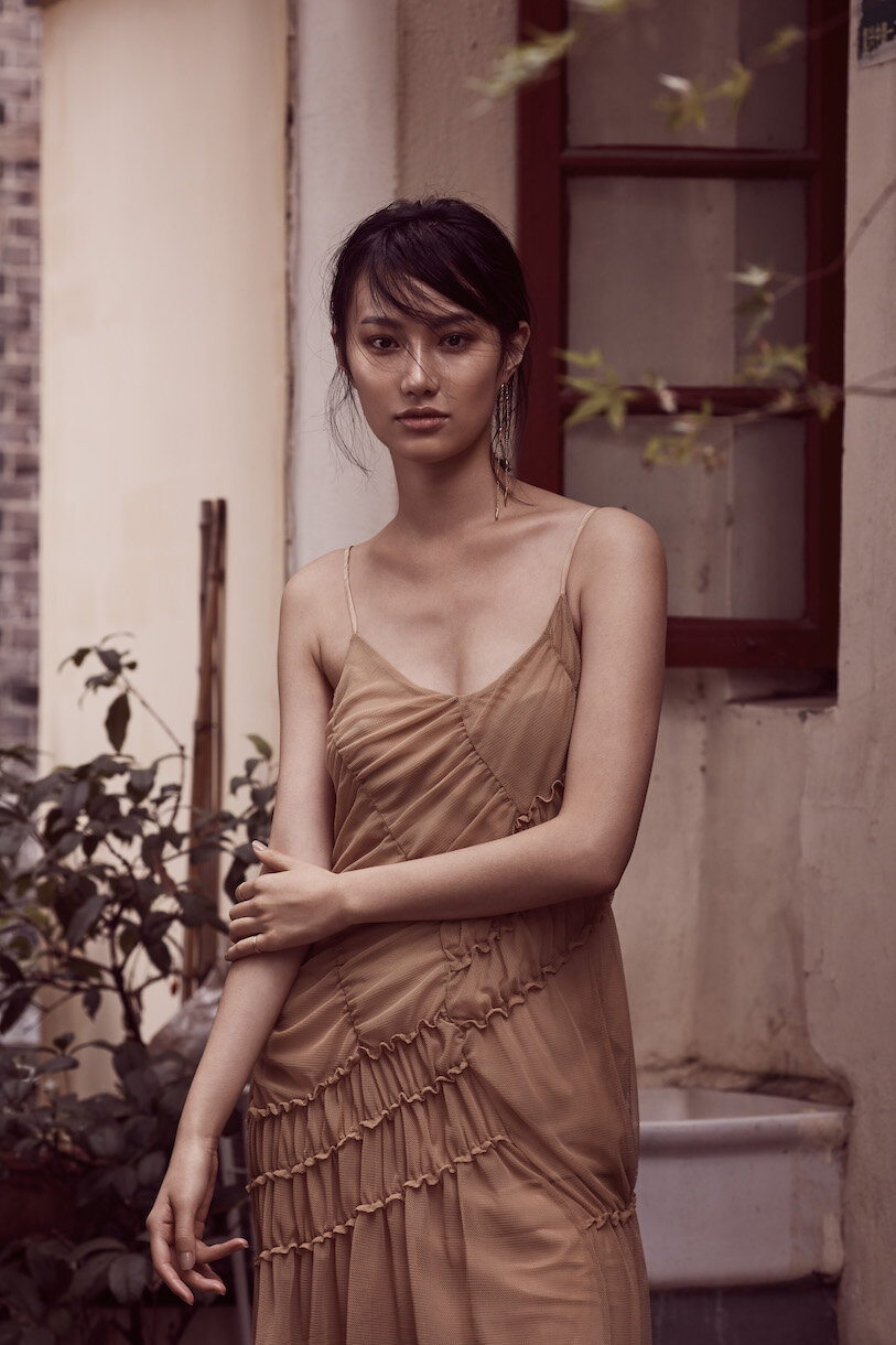 Asian woman in a flowing brown dress posing outdoors against a weathered building with green foliage; fashion editorial portrait.