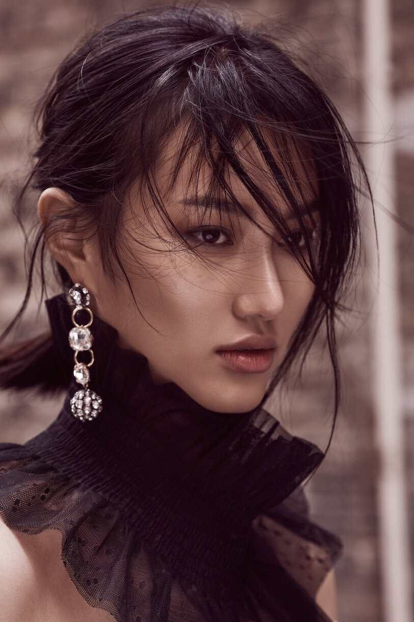 Close-up portrait of an Asian woman with dark hair, wearing a black ruffled top and long, jeweled earrings against a neutral backdrop.