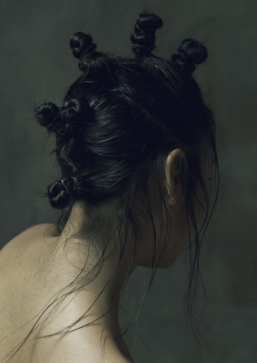 Back view of a young woman with dark hair styled in twisted knots, bare shoulders, against a dark backdrop, emphasizing hair and skin tones.