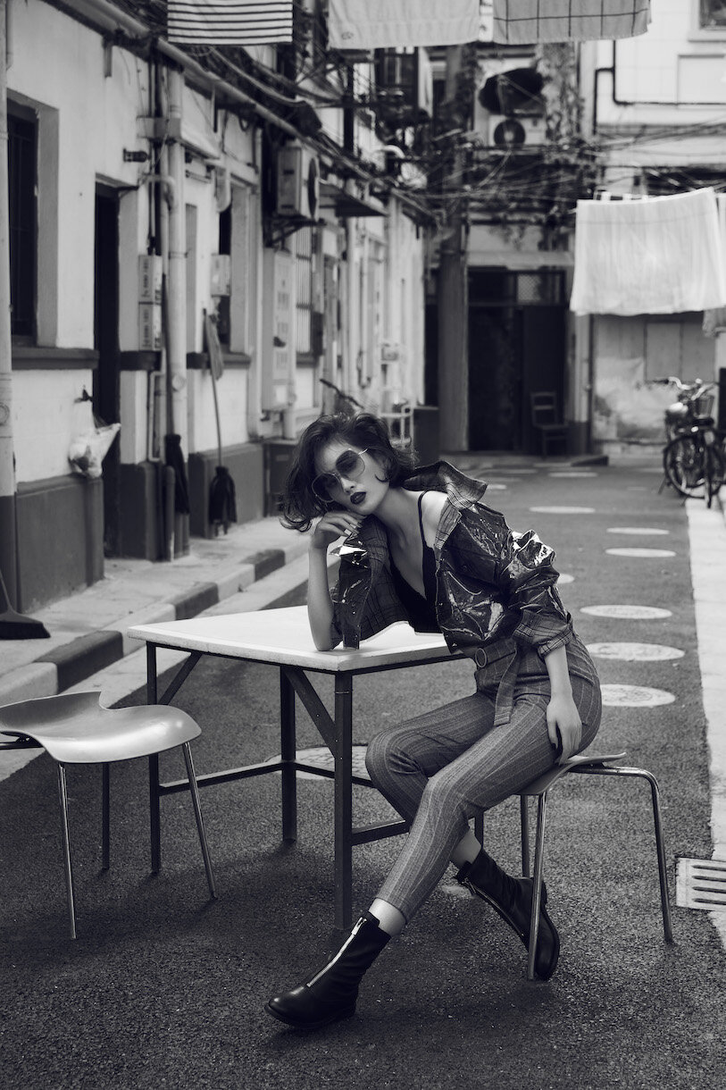 Black and white fashion shot of a woman in sunglasses, jacket, and striped pants sitting at a table in an urban alleyway.