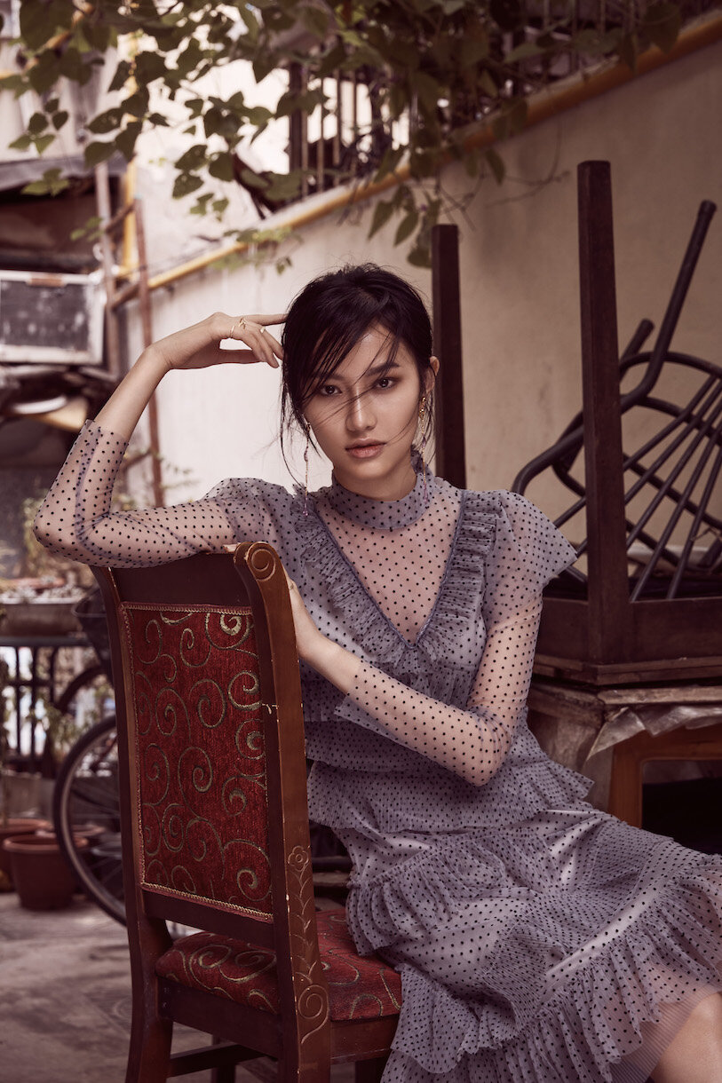 Attractive Asian woman in a dotted sheer dress sits on a vintage chair outside with plants and an old bicycle in the background.