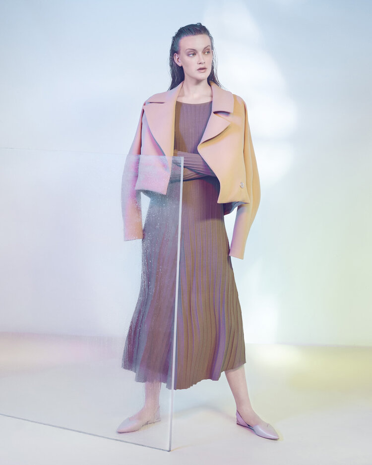 Elegant female fashion model poses in a lilac jacket and pleated midi dress against a studio backdrop with light and shadow effects.