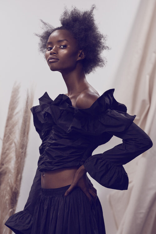 Fashion shot of a young Black woman wearing a ruffled black top and skirt, posing in front of a cream-colored draped backdrop. Studio lighting.
