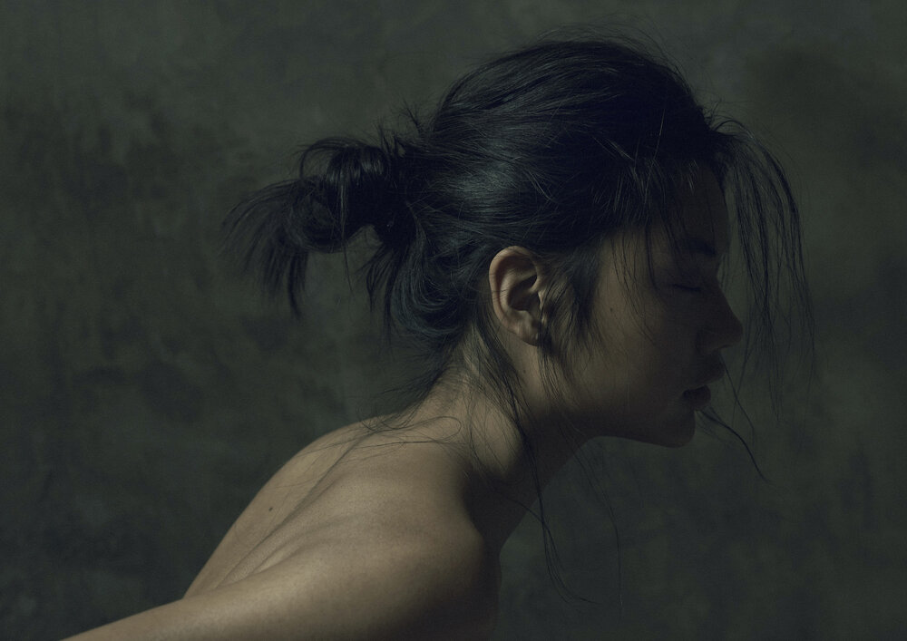Side profile portrait of a woman with dark hair in a bun against a dark textured backdrop in a dimly lit setting.