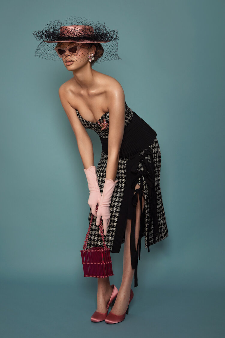 A stylish woman in a strapless dress with gloves and hat, carrying a boxy handbag, poses against a teal background in a studio shot.
