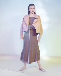 Model standing behind a glass pane wearing a pastel pink jacket and a pleated brown dress in a brightly lit studio