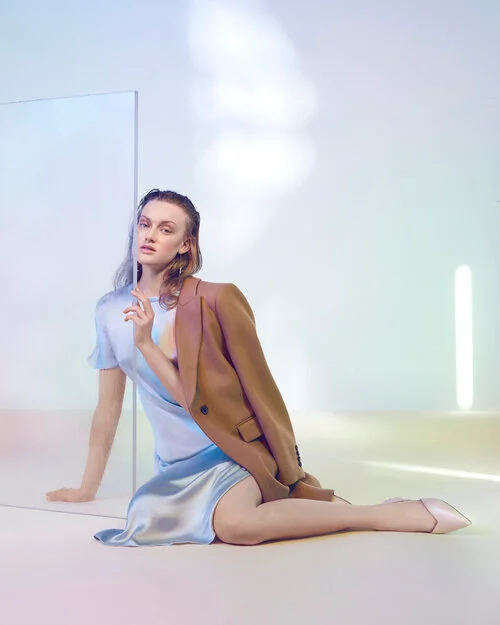 Model seated wearing a light blue satin dress and brown blazer, posed behind a clear glass partition.