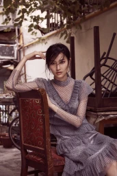 Woman in grey polka dot sheer dress seated on a wooden chair with red patterned upholstery outdoors
