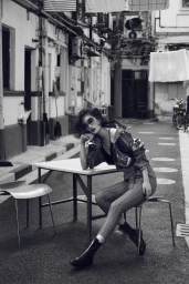 Monochrome image of a woman with short dark hair in sunglasses seated at an outdoor table in a narrow alleyway.