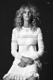 Monochrome portrait of a woman with voluminous blonde curls wearing a white patterned long-sleeve dress with hands clasped.