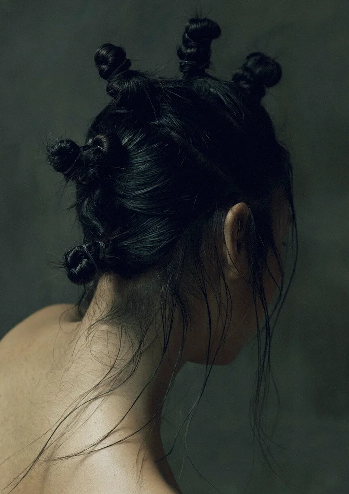 Close-up back view of a person with glossy dark hair styled into multiple small knots, set against a muted dark textured background