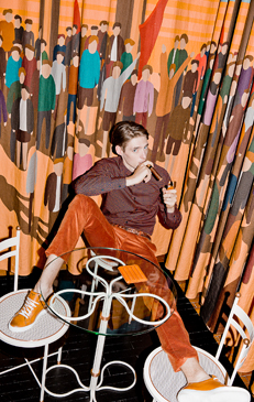 Man lighting a cigar sitting at an outdoor cafe with orange patterned curtains, wearing an orange suit and brown shirt.