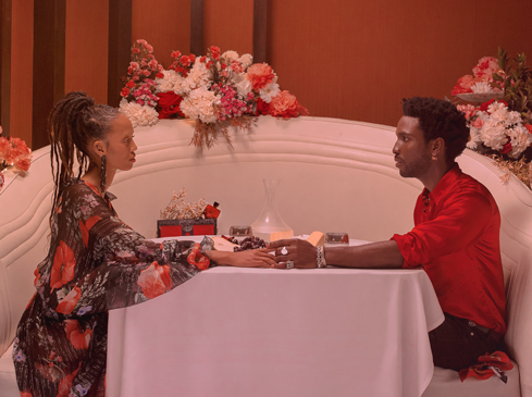 An elegant couple sits at a table with flowers and candles in a warmly lit indoor setting, hands touching, exuding romance and intimacy.