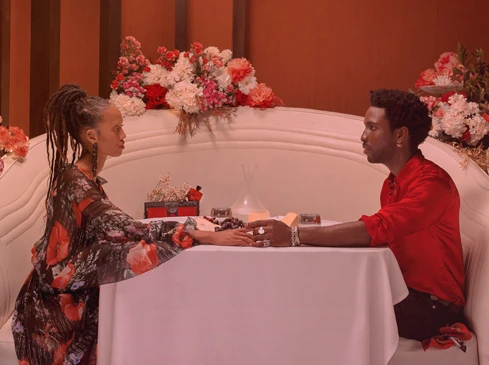 An elegant couple sits at a table with flowers and candles in a warmly lit indoor setting, hands touching, exuding romance and intimacy.