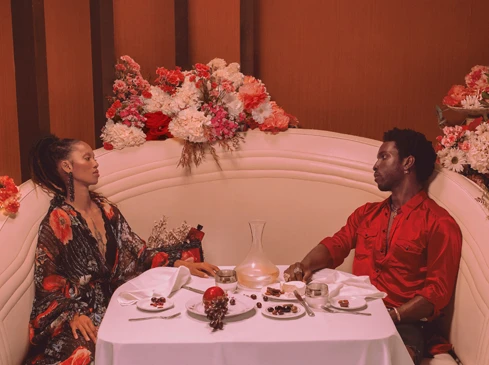 Elegant date night scene: a couple sits at a table adorned with flowers and fruit, enjoying a meal in a cozy, vintage-toned setting.