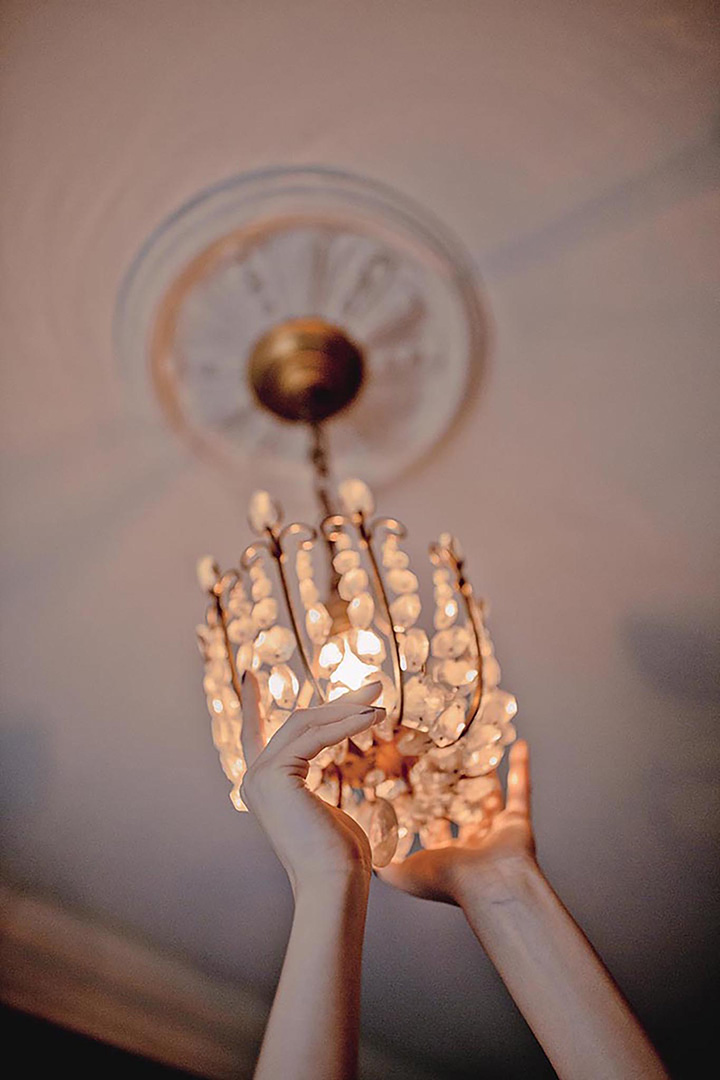 Close-up shot of a woman's hands holding a crystal chandelier against a soft pink ceiling with ornate moulding and a warm glow.