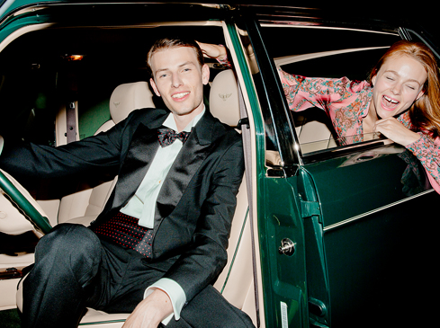 Two adults in a vintage car. The man, in a tuxedo, is in the driver's seat, while the woman leans out the back window laughing.