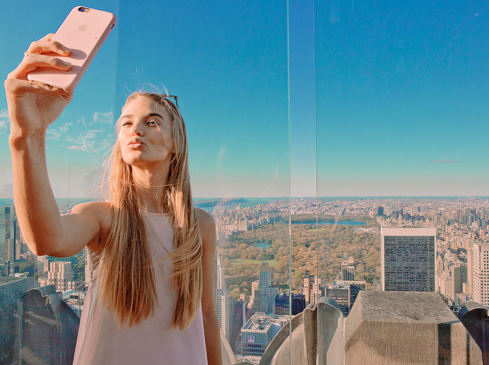 Woman takes a selfie with a pink phone on a rooftop with city skyline background, wearing a white dress in daylight.