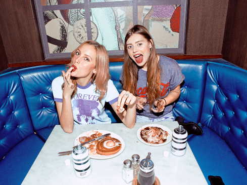 Two young women at a booth in a diner. Plates of pancakes sit on the table alongside condiments and cell phone. The booth has blue upholstery.