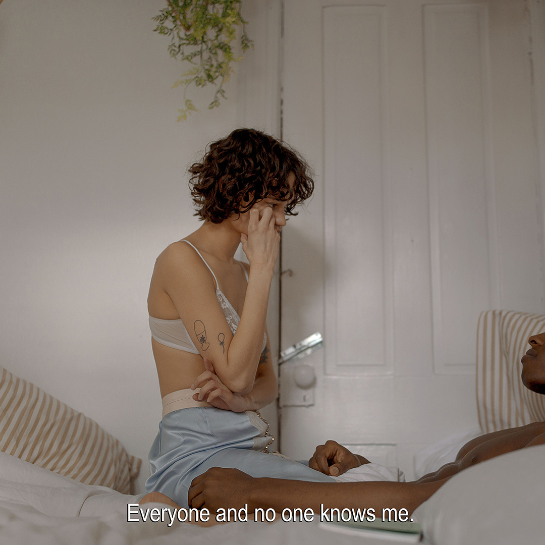 Woman sits on bed with partner in bedroom, wearing lingerie and skirt. Soft lighting illuminates the scene, highlighting the couple.