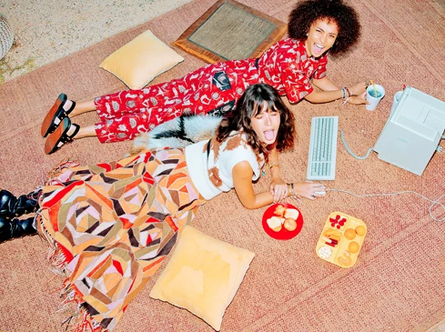 Two women lying on a rug indoors with a vintage computer, food plates, and drinks, laughing and sticking out tongue, wearing colorful retro clothing.