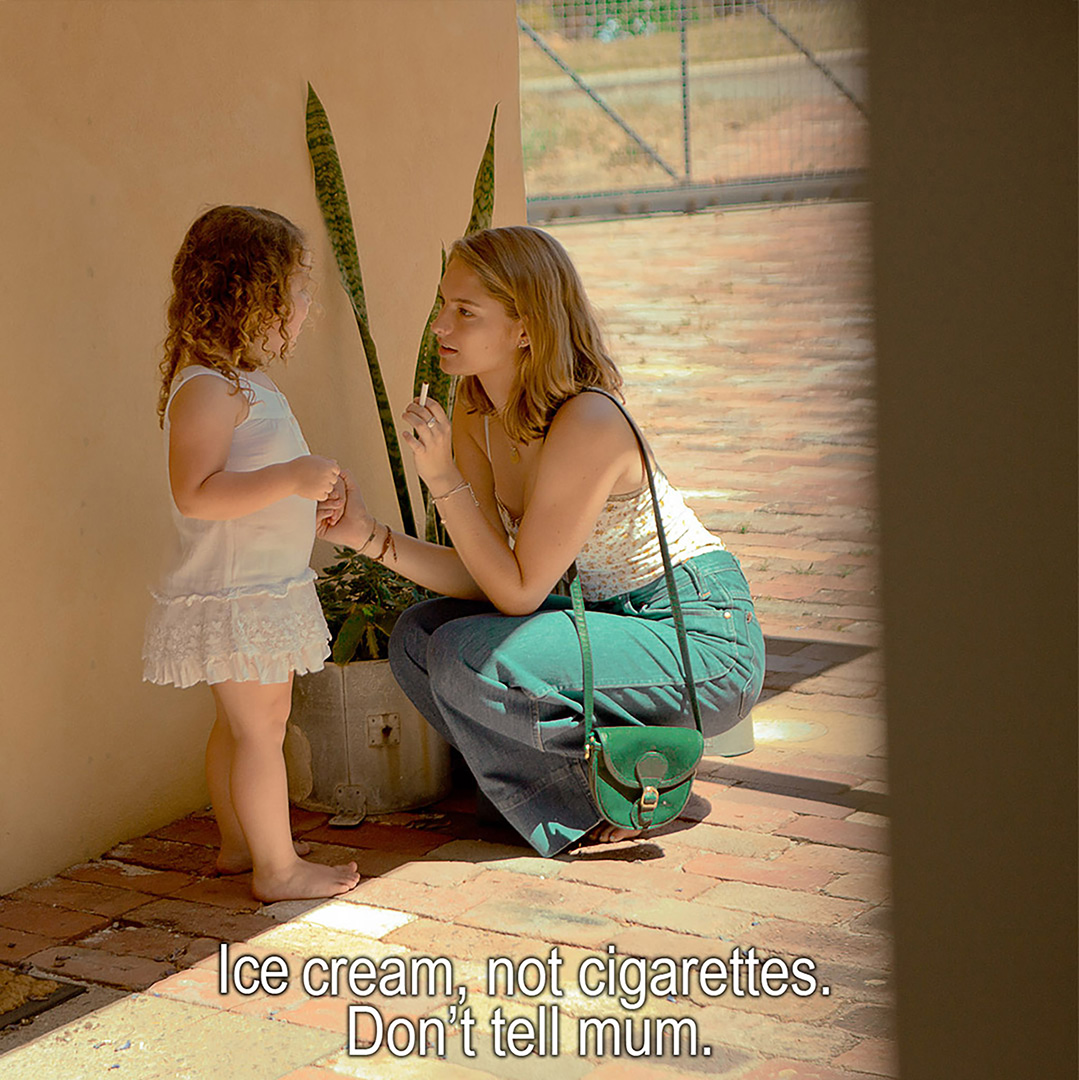A young woman squats, eye level to a little girl. They are conversing on brick patio outside. The woman is holding a thin white item.