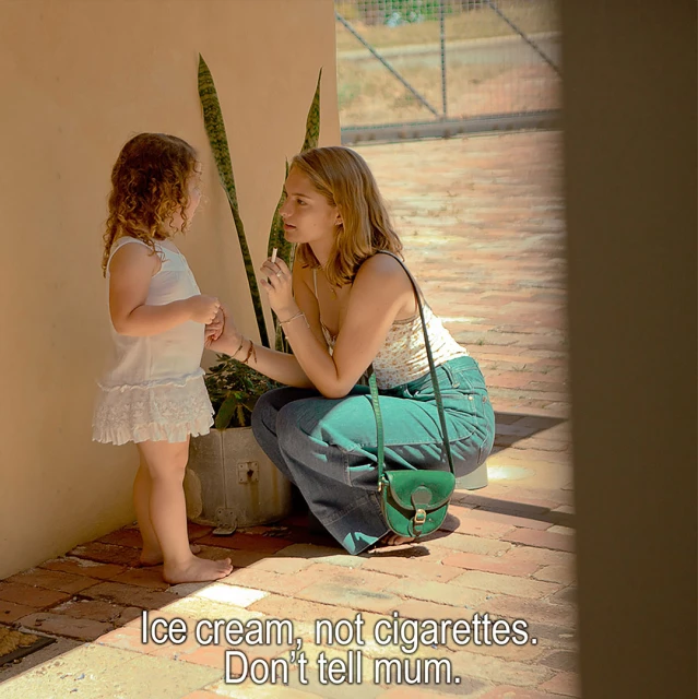 A young woman squats, eye level to a little girl. They are conversing on brick patio outside. The woman is holding a thin white item.